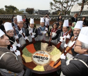 Several people wearing chef hats and posing with large wooden spoons surround a large pot of colourful dry ingredients.