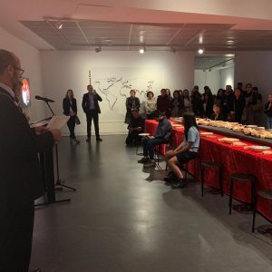 A long room with a dining table running down the centre. A presenter speaks at a microphone, and many people are gathered at the end of the table listening.