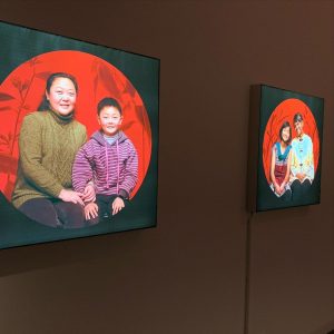 Two portraits of mothers and their children smiling with red backdrops. The portraits are lit up and placed side by side on a wall in a darkened room.