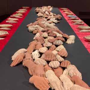 A long dining table set with a red tablecloth and black table runner. Many ceramic hands lay, overlapping in a line down the centre of the table.