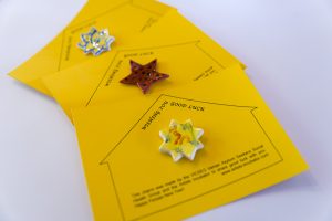 Three ceramic good luck charms. Each charm is attached to a yellow card depicting a house shape diagram with the charm attached at the centre of its house.