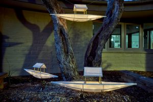 Three decorative paper boats lit up and set at the base of and upon a tree at night.