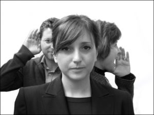Three people facing in different directions.  One person is facing forward not speaking, another is holding their hand to their ear to listen and a third person is speaking loudly.