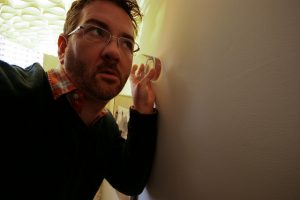 Image of a man holding a glass to the wall to try to listen to what is being said in another room.